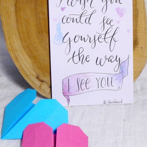 May include: A white card with black calligraphy that reads 'I wish you could see yourself the way I see you.'  The card has a watercolor banner with the words 'I see you' and a small blue beaker with a white smoke plume. The card is surrounded by pink hearts. The card is signed 'Ali Hazelwood.'  Two origami hearts, one blue and one pink, are in the foreground.