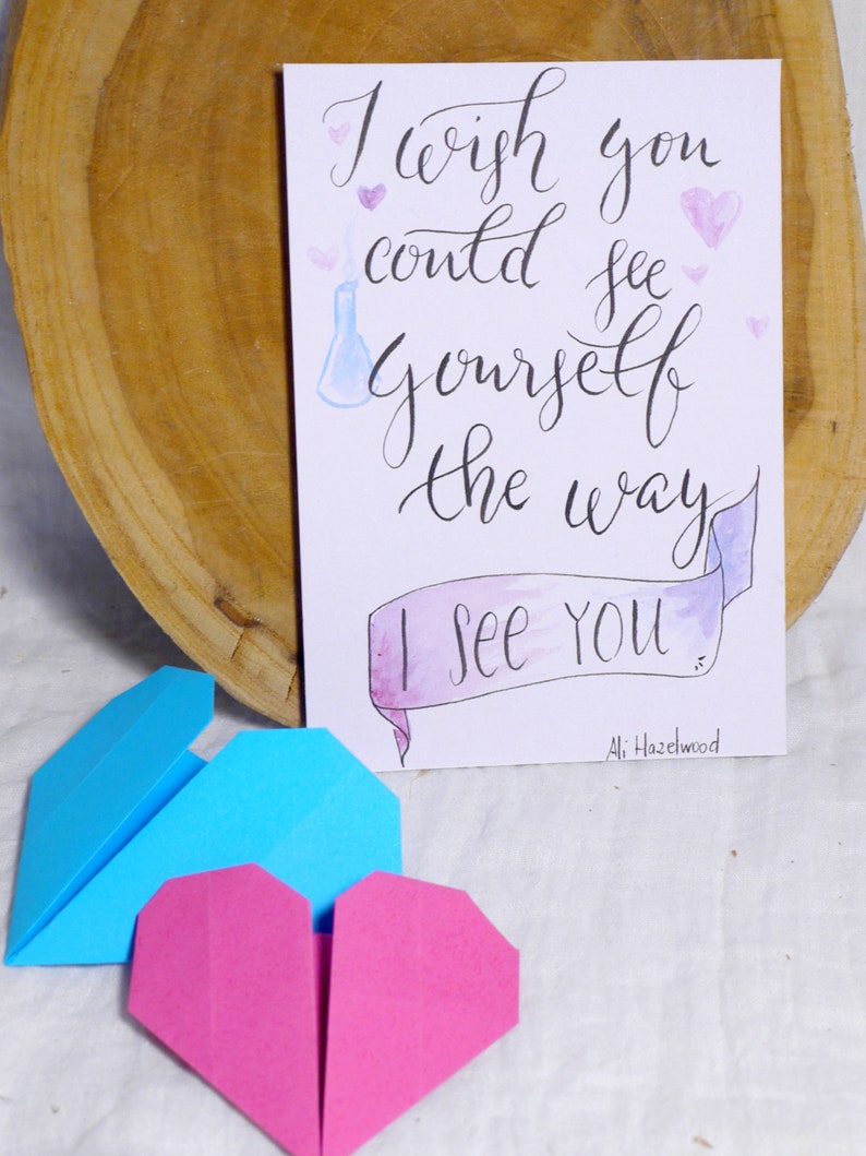May include: A white card with black calligraphy that reads 'I wish you could see yourself the way I see you.'  The card has a watercolor banner with the words 'I see you' and a small blue beaker with a white smoke plume. The card is surrounded by pink hearts. The card is signed 'Ali Hazelwood.'  Two origami hearts, one blue and one pink, are in the foreground.