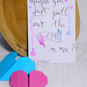 May include: A handwritten note on white paper with blue and pink watercolor washes. The note says 'maybe you just pull out the best in me?' with a pink and blue banner that says 'best' and a small blue bottle with a pink lid. The note is signed 'oli hazelwood' and there are small blue and pink hearts scattered around the note. Two origami hearts, one blue and one pink, are in the foreground.