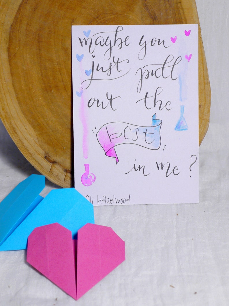 May include: A handwritten note on white paper with blue and pink watercolor washes. The note says 'maybe you just pull out the best in me?' with a pink and blue banner that says 'best' and a small blue bottle with a pink lid. The note is signed 'oli hazelwood' and there are small blue and pink hearts scattered around the note. Two origami hearts, one blue and one pink, are in the foreground.