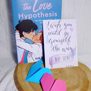May include: A blue paperback book with the title 'the Love Hypothesis' on the cover. The cover features a cartoon illustration of two people kissing. A white card with the text 'I wish you could see yourself the way I see you.' and a pink heart is next to the book. A blue origami paper airplane and a pink origami heart are on a wooden surface.