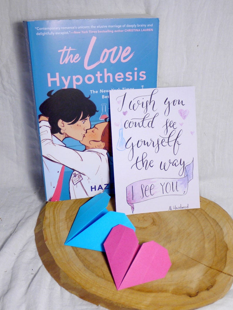 May include: A blue paperback book with the title 'the Love Hypothesis' on the cover. The cover features a cartoon illustration of two people kissing. A white card with the text 'I wish you could see yourself the way I see you.' and a pink heart is next to the book. A blue origami paper airplane and a pink origami heart are on a wooden surface.