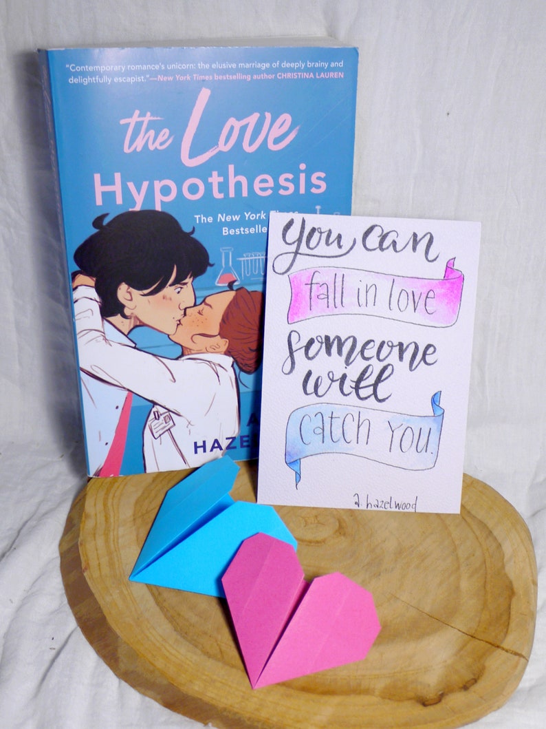 May include: A blue and pink origami heart on a wooden surface. A book titled 'The Love Hypothesis' by Ali Hazelwood is in the background. A card with the text 'You can fall in love someone will catch you' is also in the background.