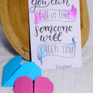 May include: A handwritten card with the text 'you can fall in love someone will catch you.' The text is written in black ink on a white background with pink and blue watercolor accents. The card is signed 'a.hazelwood.' There are two origami hearts, one blue and one pink, in the foreground.
