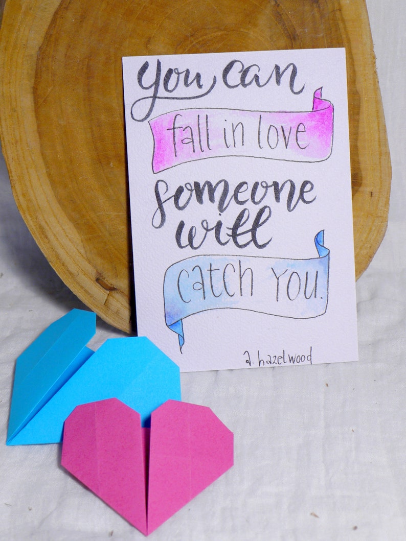 May include: A handwritten card with the text 'you can fall in love someone will catch you.' The text is written in black ink on a white background with pink and blue watercolor accents. The card is signed 'a.hazelwood.' There are two origami hearts, one blue and one pink, in the foreground.