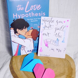 May include: A blue and pink origami heart on a wooden surface. A book titled 'The Love Hypothesis' by Ali Hazelwood is in the background. A card with the text 'maybe you just pull out the best in me?' is also in the background.