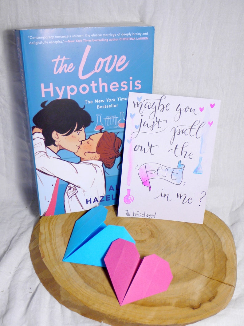 May include: A blue and pink origami heart on a wooden surface. A book titled 'The Love Hypothesis' by Ali Hazelwood is in the background. A card with the text 'maybe you just pull out the best in me?' is also in the background.