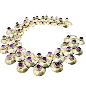 Puede incluir: Un collar plateado con una serie de elementos abovedados en forma de pétalo, cada uno con una vibrante piedra preciosa púrpura. El collar está dispuesto en forma curva sobre un fondo blanco, mostrando su diseño.