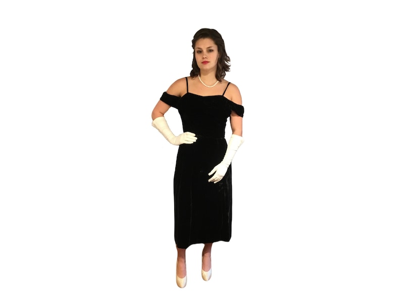 Puede incluir: Un vestido negro de terciopelo con hombros descubiertos y tirantes finos. El vestido llega a la rodilla y se combina con guantes blancos y tacones a juego. Un collar de perlas completa el look.