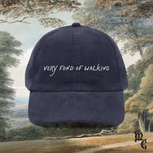 May include: Navy blue corduroy baseball cap with the embroidered text "Very Fond of Walking" in white. The cap has a curved brim and a rounded crown. The background features a landscape painting.