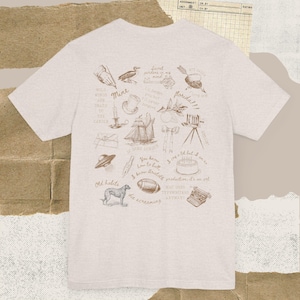 Puede incluir: Una camiseta gris claro con una serie de ilustraciones en tinta marrón. Los dibujos incluyen un barco, un perro, un pastel y frases como "Old habits" y "Florida!!!".