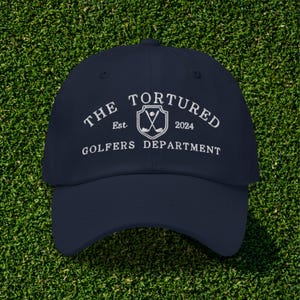 May include: A navy blue baseball cap with white embroidered text that reads "The Tortured Golfers Department Est. 2024" and a white embroidered logo of crossed golf clubs inside a shield.