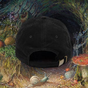 May include: Black corduroy baseball cap with a curved brim and adjustable strap. The cap features a metal buckle and a classic six-panel design. The background is a painting of a forest scene with mushrooms and a snail.