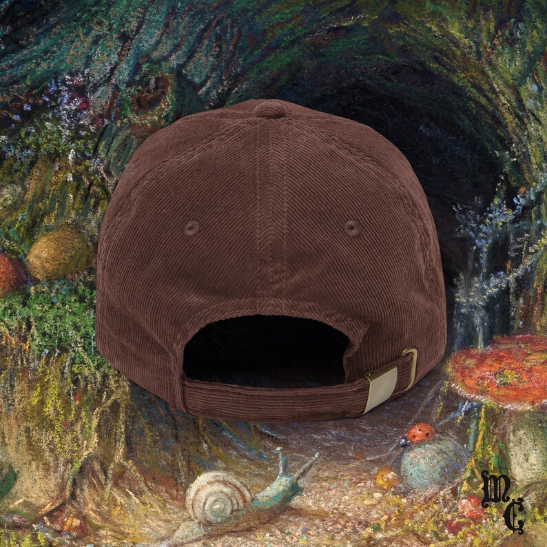 May include: A brown corduroy baseball cap with a curved brim and adjustable strap. The cap has a classic six-panel design and a metal buckle closure. The background features a colorful, whimsical illustration of a forest scene with mushrooms and a snail.