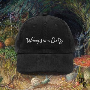 May include: Black corduroy baseball cap with the embroidered text "Whoopsie-Daisy" in white script. The cap has a curved brim and a structured crown. The background features a colorful, impressionistic painting with mushrooms and a snail.
