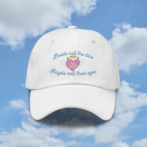 May include: White baseball cap with an embroidered design featuring a pink heart with a dice inside, angel wings, a halo, and the text "Devils roll the dice Angels roll their eyes".