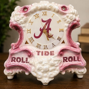 May include: A ceramic clock with a white face and gold hands, featuring Roman numerals and a maroon "A" logo. The clock has a pink and white decorative frame with the words "ROLL TIDE ROLL".
