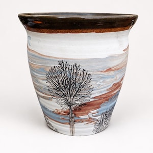 May include: A ceramic planter with a flared rim and a dark brown glazed top edge. The body features a marbled pattern of white, light blue, and earthy brown tones, overlaid with a detailed black silhouette of a tree and smaller shrubbery.