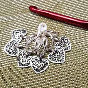May include: A set of six silver heart-shaped stitch markers with a decorative filigree design. The markers are attached to a white plastic ring. A red crochet hook is also visible in the image.