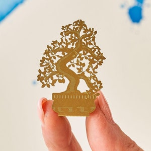 May include: A gold-colored bonsai tree-shaped metal cutout. The cutout is held between fingers against a background of blue watercolor paint and a white surface. The bonsai tree has detailed leaves and branches.