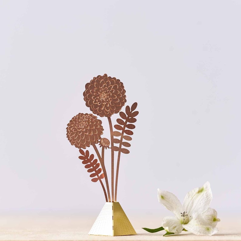 Peut inclure: Un bouquet de fleurs en papier de couleur cuivre avec trois fleurs et des feuilles, pos&eacute; sur un socle en forme de pyramide dor&eacute;e.