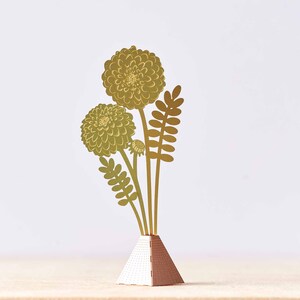 Peut inclure: Une sculpture de fleurs en m&eacute;tal dor&eacute; avec trois fleurs et des feuilles dans un vase g&eacute;om&eacute;trique dor&eacute; et blanc.