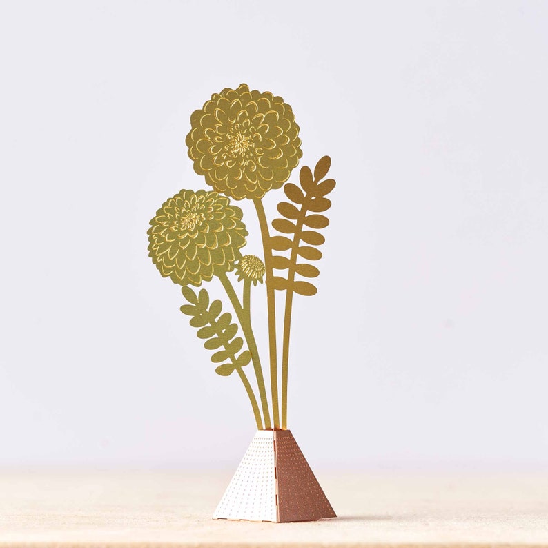 Peut inclure: Une sculpture de fleurs en m&eacute;tal dor&eacute; avec trois fleurs et des feuilles dans un vase g&eacute;om&eacute;trique dor&eacute; et blanc.