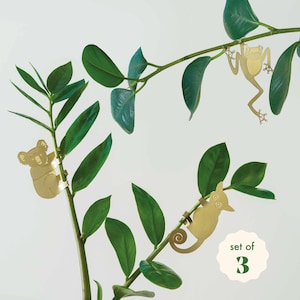 May include: Set of three brass-colored animal ornaments: a koala, a frog, and a possum. The ornaments are attached to a green leafy plant. The koala is clinging to a branch, the frog is hanging, and the possum is hanging with its tail curled.