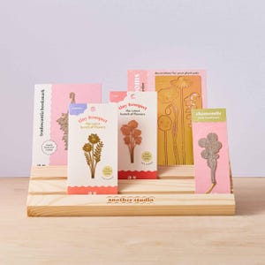 Peut inclure: Un pr&eacute;sentoir en bois avec une vari&eacute;t&eacute; de marque-pages en m&eacute;tal aux motifs floraux. Les marque-pages sont &eacute;tiquet&eacute;s avec les noms "Tradescantia", "Tiny Bouquet", "Chamomile" et "Blooms Garden".