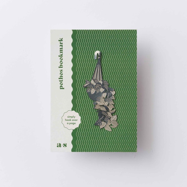 May include: A silver metal bookmark shaped like a hanging pothos plant. The bookmark has a hook at the top to hang over a page. The card reads "pothos bookmark" and "simply hook over a page".