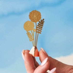 Peut inclure: Un bouquet de fleurs en papier dor&eacute; avec trois fleurs et des feuilles dans un petit vase orange en forme de pyramide.