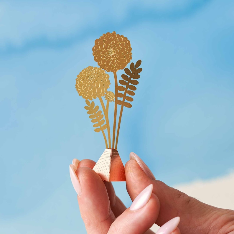 Peut inclure: Un bouquet de fleurs en papier dor&eacute; avec trois fleurs et des feuilles dans un petit vase orange en forme de pyramide.