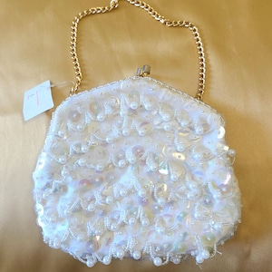May include: A white beaded handbag with a gold chain handle. The bag is covered in sequins and small pearls. A tag is attached to the handle. The bag is on a gold background.