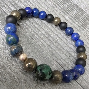 May include: A beaded bracelet featuring a mix of matte black, metallic gold, and deep blue lapis lazuli beads. The bracelet has a stretch design and is displayed on a gray wooden surface. The beads vary in size and texture.