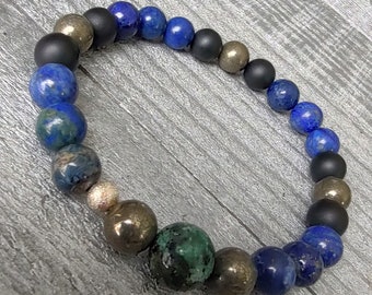 Pode incluir: Uma pulseira de contas com uma mistura de contas pretas foscas, douradas metálicas e lápis-lazúli azul escuro. A pulseira é exibida em uma superfície de madeira cinza. As contas variam em tamanho e textura.