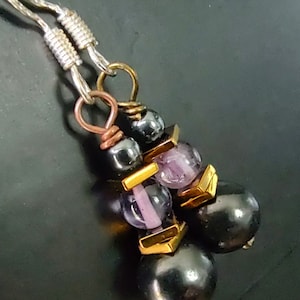 Può includere: Primo piano di orecchini fatti a mano con perline grigio scuro e viola, distanziatori quadrati color oro, ganci per le orecchie color argento e anelli color rame. Le perline sono infilate insieme per creare un effetto pendente.