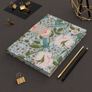 Może przedstawiać: Niebieski i różowy notes z kwiatowym wzorem i miękką, fakturowaną okładką. Notes jest zamknięty i leży na brązowej powierzchni z ołówkami i spinaczami.