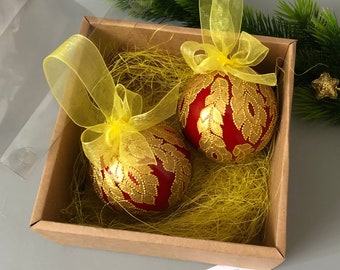 May include: Two red Christmas ornaments with gold leaf designs and yellow ribbon bows are displayed in a brown box with yellow shredded paper. The box is open and the ornaments are visible. A business card for "Pin of Color" is in the foreground.