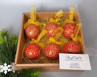 May include: Six red Christmas ornaments with gold painted designs are displayed in a box with yellow tissue paper. The ornaments are ready to be hung on a Christmas tree.
