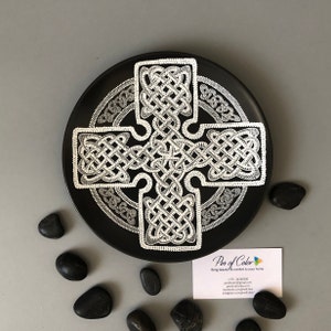 Könnte beinhalten: Ein schwarzer Keramikteller mit einem weißen keltischen Knotendesign. Der Teller ist rund und hat einen erhöhten Rand. Das Design ist kompliziert und detailliert und befindet sich in der Mitte des Tellers. Der Teller ist von einem weißen Rand umgeben.