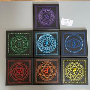 May include: Seven black square canvases with colorful mandala designs representing the seven chakras. Each canvas features a different color and symbol, including Om, Muladhara, Svadhisthana, Manipura, Anahata, Vishuddha, and Ajna.