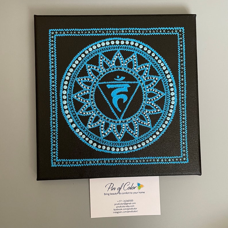 May include: A blue and white mandala painting on a black canvas. The mandala features a stylized symbol with a triangle and a circle. The painting is framed with a black border.
