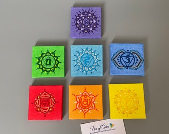 May include: Seven small square canvases painted with the seven chakra symbols in their corresponding colors. The canvases are arranged in a rainbow pattern from top to bottom, starting with purple and ending with yellow. Each canvas features a mandala design with the chakra symbol in the center. The canvases are all the same size and are approximately 4 inches by 4 inches.