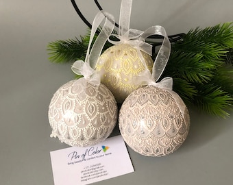 May include: Three hand-painted Christmas ornaments with a white and silver design. The ornaments are round and have a delicate pattern. The ornaments are hanging from white ribbon.
