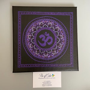 May include: Purple and white mandala with the Om symbol in the center, painted on a black canvas. The canvas is framed with a purple border.