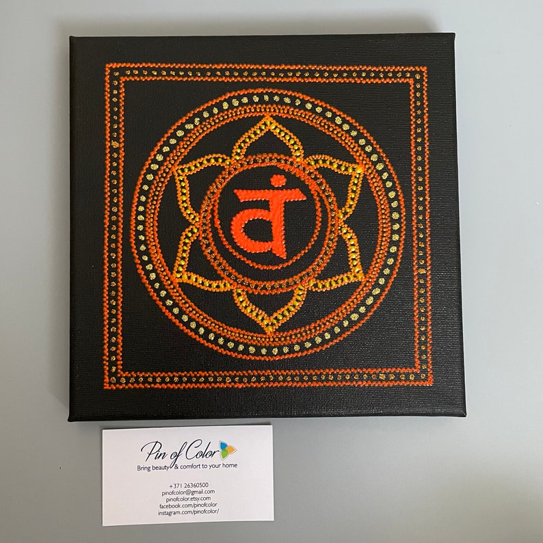 May include: A square canvas painting with a black background and a large orange mandala with a Sanskrit symbol in the center. The mandala is surrounded by a gold border.