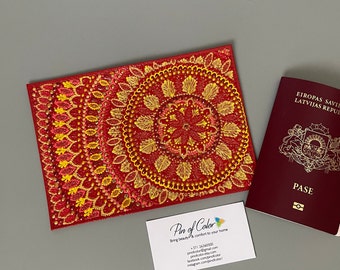 May include: Red passport cover with a yellow and orange mandala design. The cover is made with paint and beads. The text "Pin of Color" is on a business card below the cover.