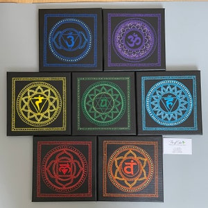 May include: Seven square canvases with black backgrounds and colorful mandala designs. Each canvas features a different color and symbol representing the seven chakras. The canvases are arranged in a row, with the top canvas featuring a purple mandala with the symbol "Om" in the center.