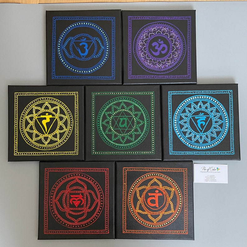 May include: Seven square canvases with black backgrounds and colorful mandala designs. Each canvas features a different color and symbol representing the seven chakras. The canvases are arranged in a row, with the top canvas featuring a purple mandala with the symbol "Om" in the center.