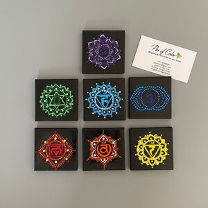 May include: Seven small black canvas paintings with colourful mandala designs representing the seven chakras. Each mandala features a different colour and symbol. The paintings are arranged in a row, with the first painting on the left and the last painting on the right.